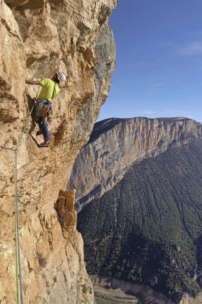 Un escalador de clásica progresa por una pared de roca caliza con un rack completo de friends y fisureros; al fondo se aprecia una gran pared y un valle profundo bajo un cielo azul.
