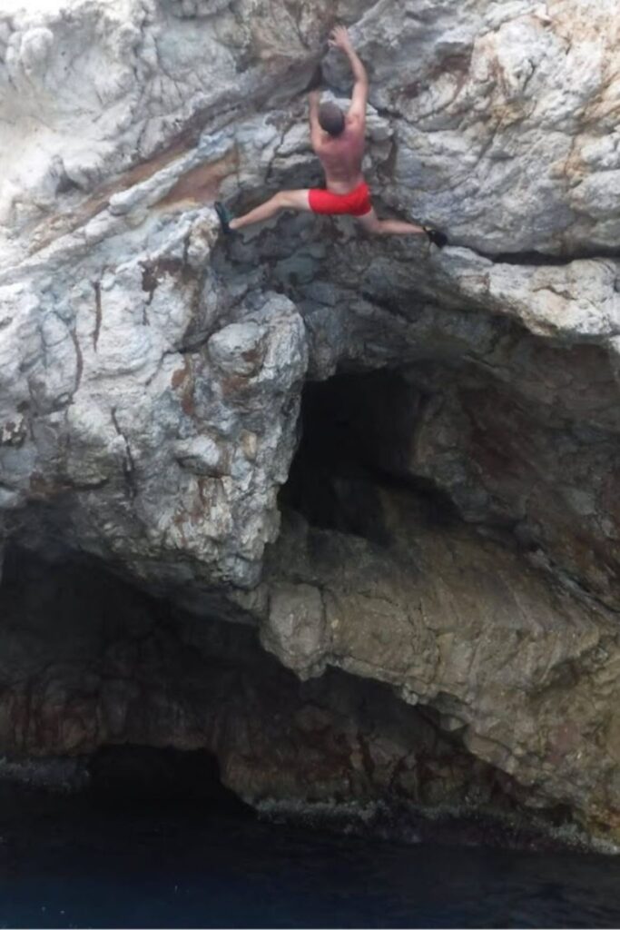 Un escalador realiza un movimiento dinámico de psicobloc sobre el mar en un acantilado extraplomado; se aprecia la altura y la zona de caída libre hacia el agua azul