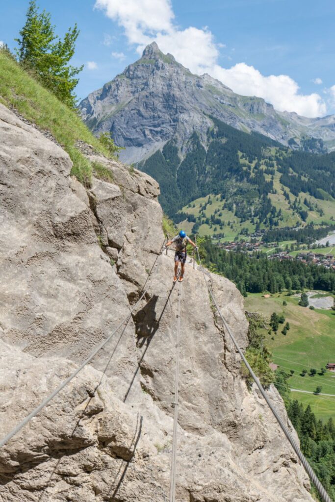 Un escalador progresa con seguridad por un puente tibetano en una **vía ferrata**, manteniendo el equilibrio sobre el cable inferior mientras está anclado con el disipador de energía a los cables de vida superiores.