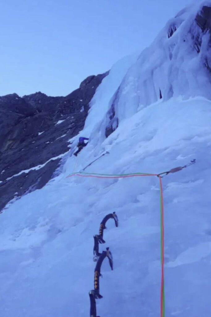 Perspectiva desde una reunión de escalada en hielo donde se ven dos piolets técnicos clavados, cuerdas dobles de colores y un escalador progresando por una cascada de hielo vertical bajo un cielo crepuscular.